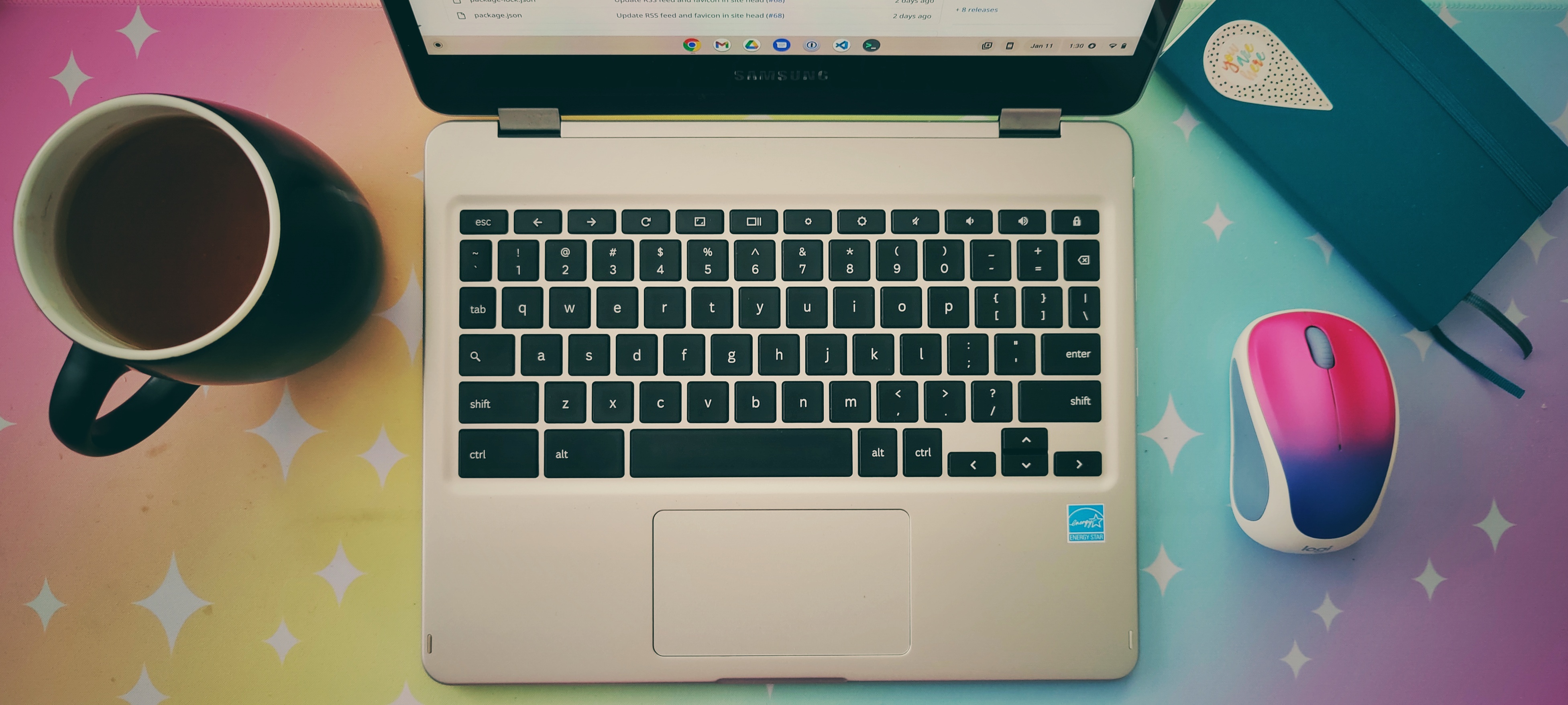 Arial view of a desktop, with an open Chromebook in the middle. A cup of tea is on the left of the Chromebook. On the right is a small blue notebook and a compact two-button mouse. Underneath is a desk mat, a rainbow pastel gradient with stars on it.